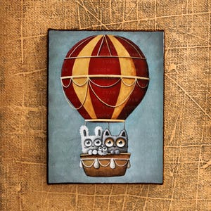 May include: A painting of three cartoon animals, a cat and two rabbits, riding in a hot air balloon. The balloon is red and yellow with white stripes and has a basket with white trim. The background is a light blue.