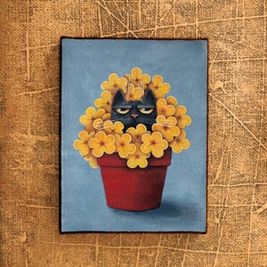 Op de afbeelding: Een zwarte kat met een chagrijnige uitdrukking verstopt zich in een pot met gele bloemen. De pot is rood en heeft een blauwe achtergrond.