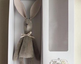 May include: A gray plush bunny rabbit toy with long ears and long legs. The bunny is wearing a light brown ribbon around its neck. The bunny is in a white box with a clear window. The box has the text "Mini Me's handmade dolls" printed on it.