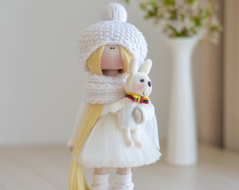 May include: A white doll with blonde hair wearing a white knitted hat, scarf, and dress. The doll is holding a white stuffed bunny rabbit.