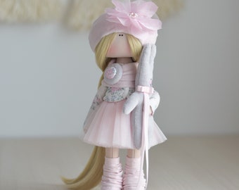 May include: A handmade doll with blonde hair, wearing a pink dress, a pink hat with a flower, and pink boots. The doll is holding a gray stuffed bunny.