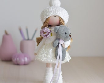 May include: A white fabric doll with blonde hair wearing a white knitted hat and a white dress with lace trim. The doll is holding a gray teddy bear with a purple ribbon.
