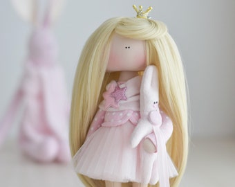 May include: A handmade doll with long blonde hair, wearing a pink dress with white polka dots and a pink star on the chest. The doll is holding a white bunny and wearing a gold crown.