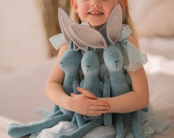 May include: A young girl wearing a blue headband with feathers holds three blue plush bunny toys. She is wearing a light blue tulle dress.