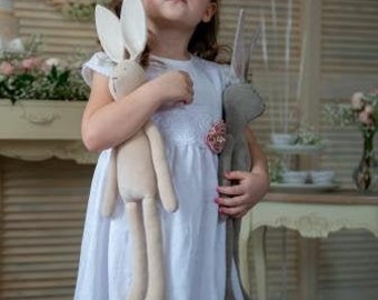 May include: A young girl in a white dress holds two plush bunny toys. The bunnies are both standing upright and have long ears. The girl is looking up and smiling.