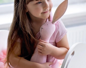 May include: A young girl wearing a pink and white flower headband with a pink bunny plush toy.