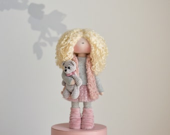 May include: A handmade doll with blonde curly hair wearing a pink and grey outfit and holding a grey teddy bear. The doll is standing on a pink quilted box.