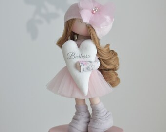 May include: A pink fabric doll with brown hair, wearing a pink hat with a flower, a pink tutu, and grey socks. The doll is holding a white heart-shaped pillow with the name "Barbara" embroidered on it. The doll is standing on a pink quilted box.