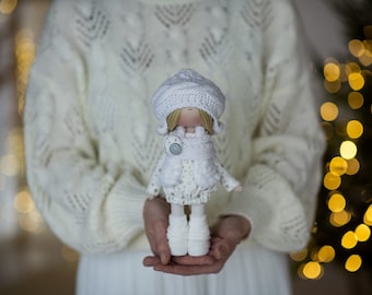 May include: A white knitted doll wearing a white knitted hat and a white knitted scarf. The doll is holding a white pom-pom in its hand. The doll is being held by a person wearing a white sweater.