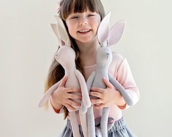 May include: A young girl with long brown hair smiles while holding two plush bunny toys. The bunnies are light pink and gray with long ears and long bodies. The girl is wearing a pink long-sleeved shirt and a gray tulle skirt.