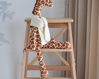 May include: A plush giraffe toy with brown and white patterned spots, wearing a cream-colored knitted scarf, sits on a wooden step stool. The giraffe's long neck and legs are visible, and the background features a light-colored wall and curtains.