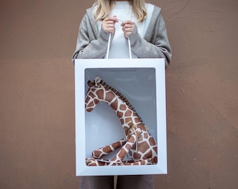 May include: A white gift bag with a clear window, featuring a brown and white stuffed giraffe inside. The giraffe is sitting with its head turned to the side.