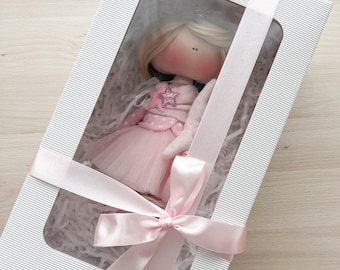 May include: A white gift box with a pink ribbon and a doll inside. The doll is wearing a pink dress with white polka dots and a star on the chest. The box has a clear window to see the doll inside.