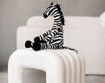 May include: A white plush stool with a rounded, arch-shaped design. A black and white zebra stuffed animal sits on top of the stool.