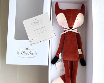 May include: A plush fox toy in a white box. The fox is brown with a white belly and a white tail tip. The fox has black eyes and a black nose. The box has a white background with the text "Mini Me" in gold lettering.