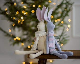 May include: Two plush bunny dolls, one cream and one gray, sit side-by-side on a wooden bench. The cream bunny wears a white scarf, and the gray bunny wears a pink and gray striped scarf. The background features a blurred Christmas tree with bokeh lights.