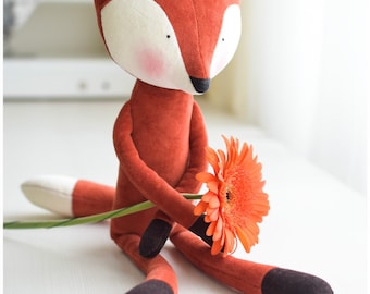 May include: A plush fox toy with a white face and orange fur is sitting on a white surface. The fox is holding a single orange gerbera daisy.