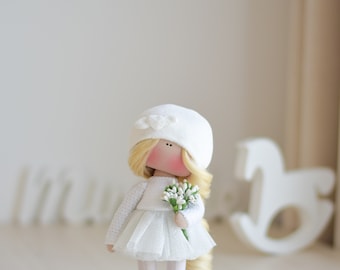 May include: A handmade doll dressed in a white dress, hat, and shoes, holding a bouquet of white flowers. The doll has blonde hair and a rosy complexion. A white rocking horse is in the background.