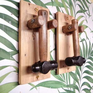 May include: Two wall-mounted hooks made from repurposed wooden hammer handles and black metal hammer heads. Each handle is attached to a rectangular light-coloured wooden plaque with visible wood grain, secured by gold-toned metal screws.