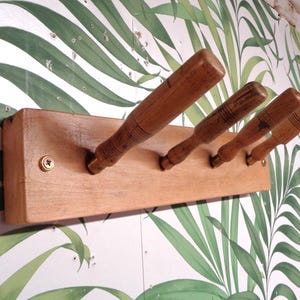 May include: A wooden coat rack with four pegs. The pegs are made of wood and are angled upwards. The rack is mounted on a light brown wooden board. The background features a white wall with a green leaf pattern.