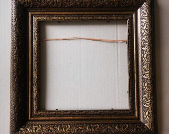 May include: Square picture frame with ornate, embossed detailing. The frame is a dark brown with gold accents, featuring intricate scrollwork along the edges. The frame is empty, with a neutral background visible through the opening.