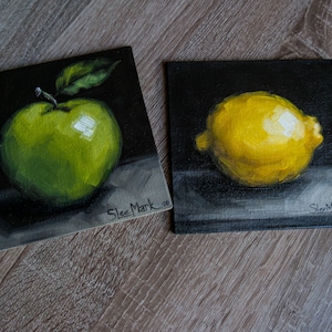May include: Two small paintings on black canvases, one depicting a green apple and the other a yellow lemon. Both paintings are signed "Slez Mark"