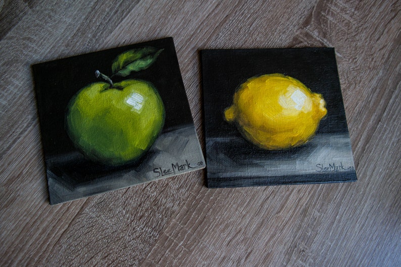 May include: Two small paintings on black canvases, one depicting a green apple and the other a yellow lemon. Both paintings are signed "Slez Mark"