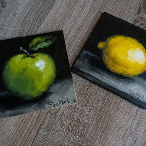 May include: Two small paintings on black canvases, one with a green apple and the other with a yellow lemon. Both paintings are signed "Slee Mark".