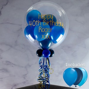 May include: A clear bubble balloon filled with blue and navy balloons. The balloon has the text "Happy 40th Birthday Roger XXX" written on it in gold lettering. The balloon is tied with blue and gold ribbon. The image also shows three additional balloons, two blue and one navy.