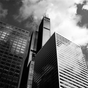Puede incluir: Foto en blanco y negro de tres edificios altos en una ciudad. El edificio más alto está en el centro de la imagen y tiene una punta puntiaguda. Los otros dos edificios están a cada lado del edificio más alto. El cielo está nublado.