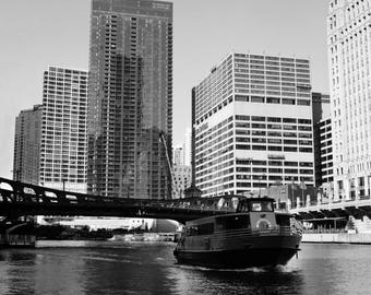 May include: A black and white photo of a boat on a river in front of a city skyline. The boat is a tour boat with a large window and a sign that says "Chicago's First Lady". The city skyline is made up of tall buildings with many windows.