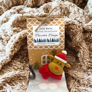 May include: A festive car diffuser gift set featuring a reindeer figurine in a red scarf and hat. The packaging is a gingerbread house design with the text "Gem Belle Festive Car Diffuser Chocolate Orange Handmade in Yorkshire".