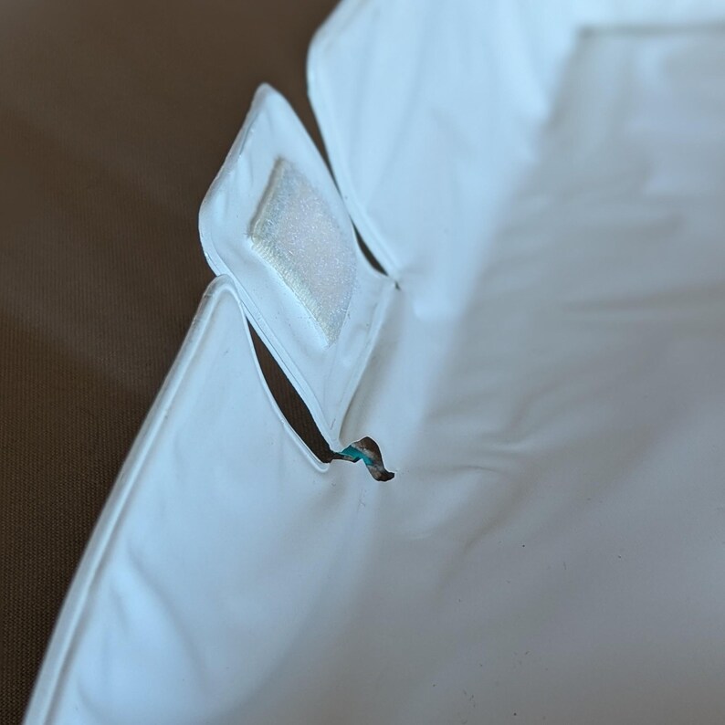 May include: A close-up of a white, waterproof, rectangular item with a velcro closure. The item has a curved edge and a small tear. The background is a solid brown color.