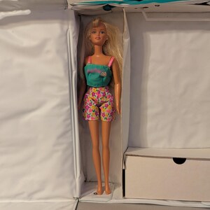 May include: A blonde doll wearing a teal top with a flower design and pink straps, paired with floral print shorts. The doll is inside a white box, suggesting a toy or collectible item.