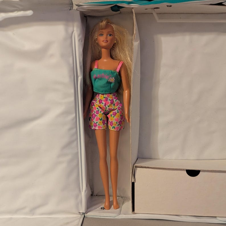 May include: A blonde doll wearing a teal top with a flower design and pink straps, paired with floral print shorts. The doll is inside a white box, suggesting a toy or collectible item.