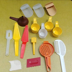 May include: A collection of 14 yellow, orange, white, and brown plastic kitchen utensils, including measuring cups, a funnel, a spatula, a grater, and a pastry cutter. The utensils are arranged on a yellow table.