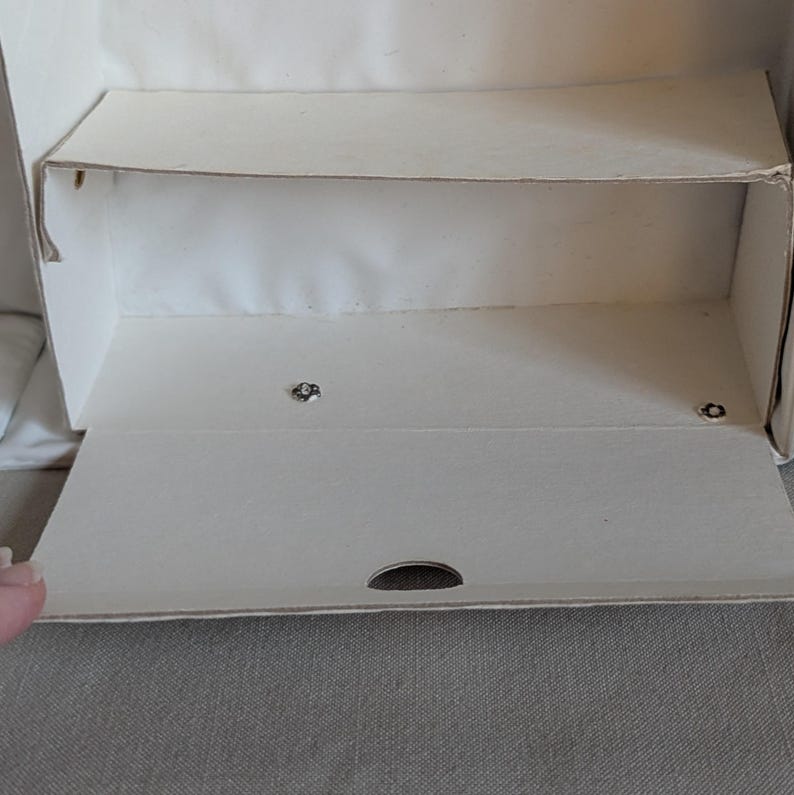 May include: An open, empty, off-white cardboard box with a hinged lid and a shelf. The lid is partially open, revealing the interior. The box has a small, oval-shaped opening on the front. The box is sitting on a gray surface.