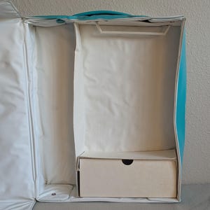 May include: An open, empty turquoise and white garment bag with a handle. The interior has a compartment with a hanger bar and a small, rectangular box at the bottom. The bag is designed to store and transport clothing.