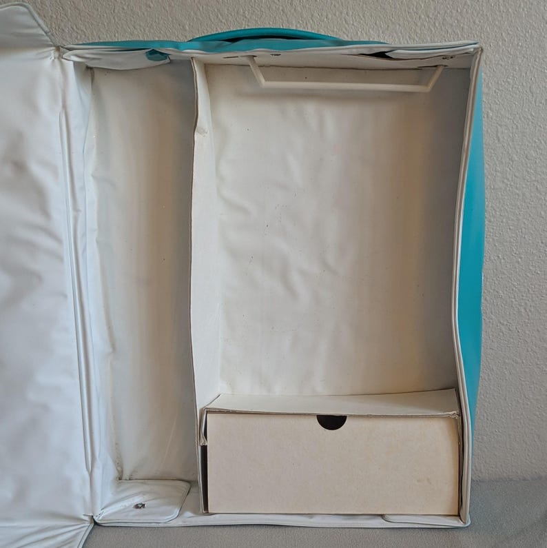 May include: An open, empty turquoise and white garment bag with a handle. The interior has a compartment with a hanger bar and a small, rectangular box at the bottom. The bag is designed to store and transport clothing.