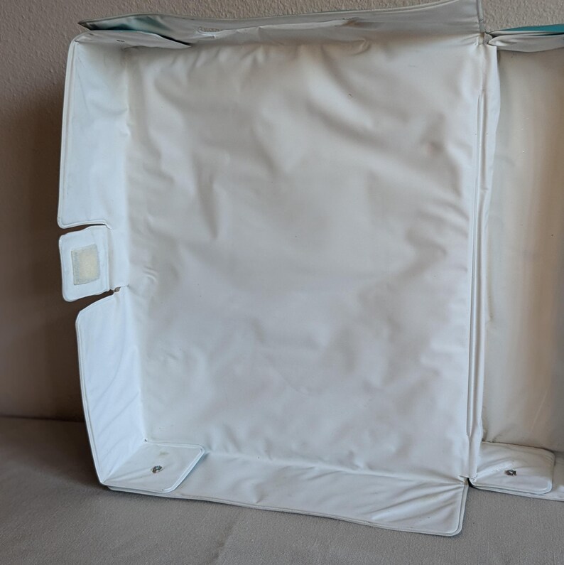 May include: White, rectangular baby changing pad with a waterproof surface. The pad has a raised edge and a velcro closure. The pad is designed to be folded for storage and transport. The pad is made of a soft, durable material.