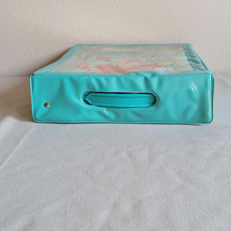 May include: A teal-colored carrying case with a clear top, featuring a handle and a gold-colored rivet. The top displays an illustration of characters from "The Little Mermaid". The case is rectangular and appears to be made of a vinyl-like material.