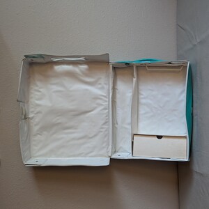 May include: An open, empty, white and teal travel case with a handle. The interior is lined with white fabric and includes a small, light-colored drawer. The case is designed to hold items for travel.