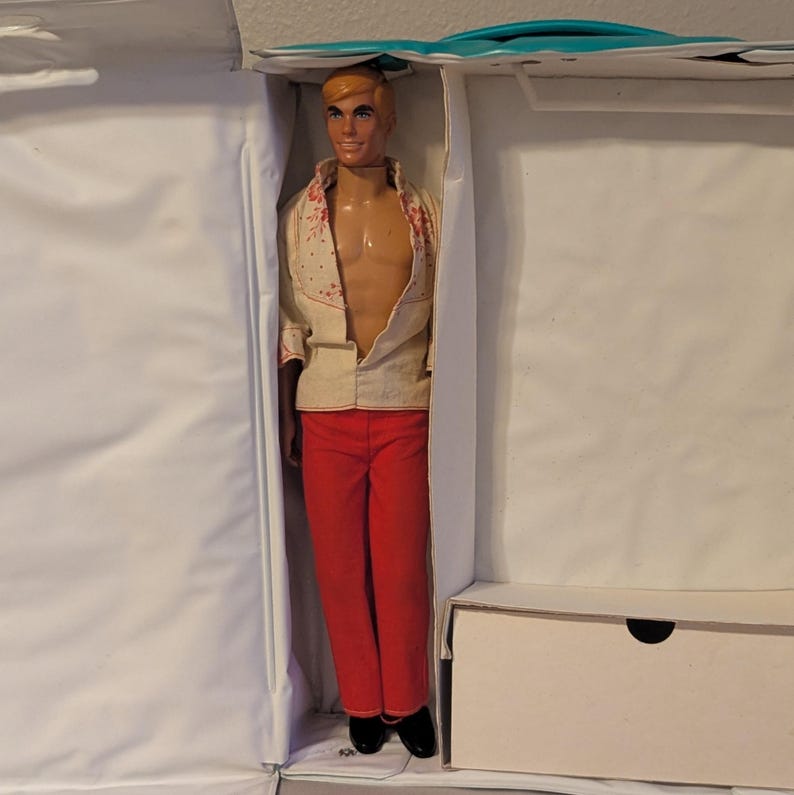May include: A vintage doll with blonde hair, wearing a cream-colored shirt with red floral accents, red pants, and black shoes. The doll is displayed inside a white box with a turquoise handle.