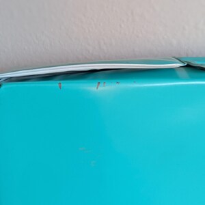 May include: A close-up of a section of a turquoise object with a white trim. The object has some minor imperfections. The background is a plain, off-white wall.