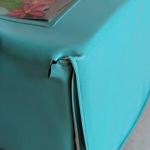 May include: Close-up of a turquoise metal box with a floral design on the lid. The box has a dented corner, revealing the metal underneath. The box is likely vintage or antique.