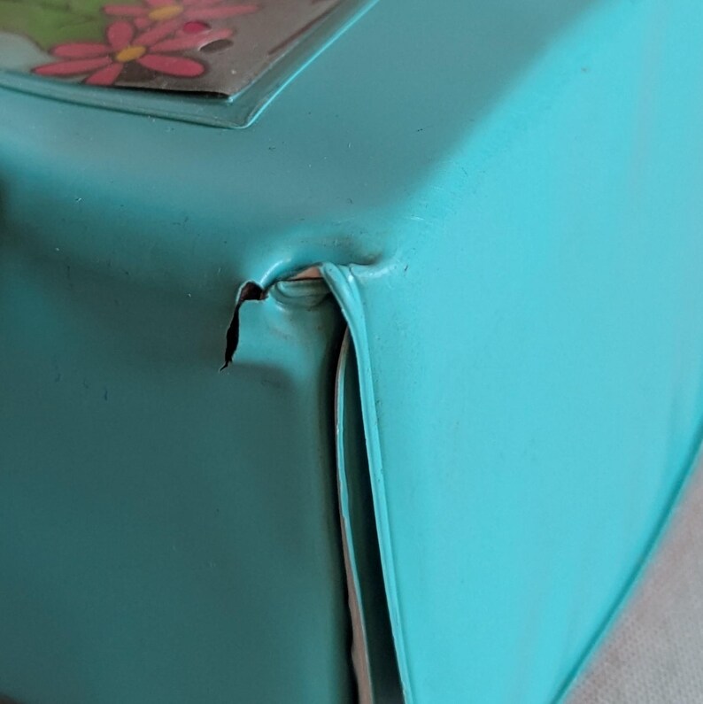 May include: Close-up of a turquoise metal box with a floral design on the lid. The box has a dented corner, revealing the metal underneath. The box is likely vintage or antique.