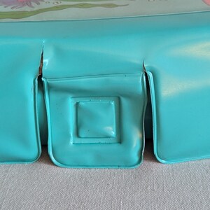 May include: Close-up of a section of a vintage turquoise metal lunchbox. The lunchbox has a rectangular shape with a square indentation and a seam. The metal has some wear and tear.