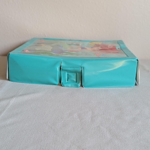May include: A rectangular, turquoise-colored box with a clear plastic top. The top features a colorful illustration. The box has a square clasp on the front. The box is sitting on a white surface.
