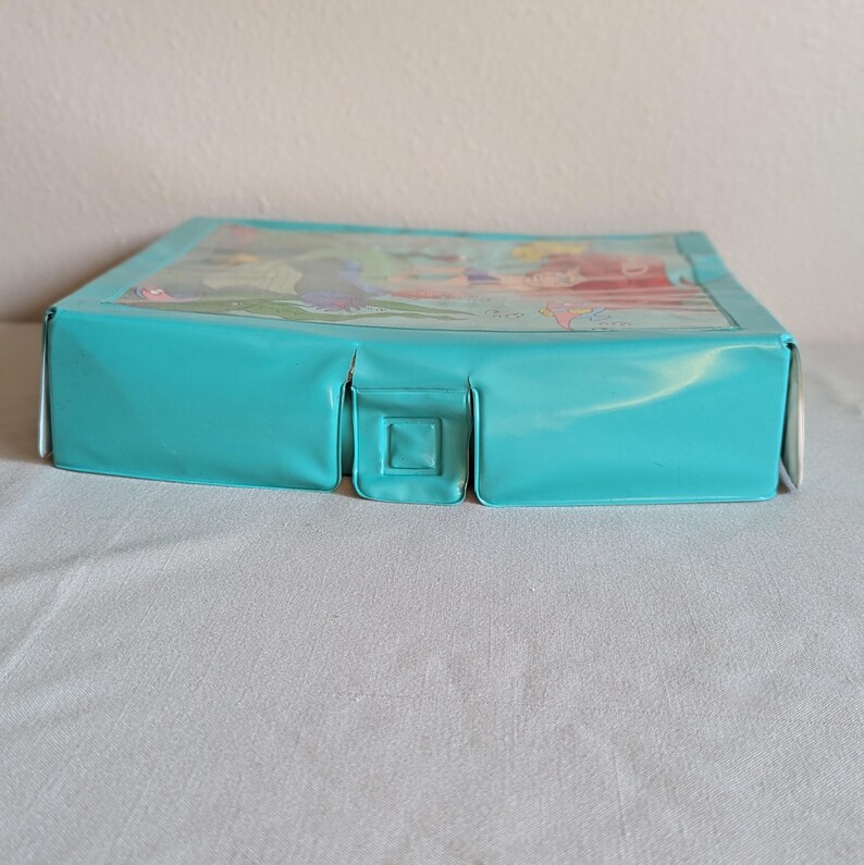 May include: A rectangular, turquoise-colored box with a clear plastic top. The top features a colorful illustration. The box has a square clasp on the front. The box is sitting on a white surface.