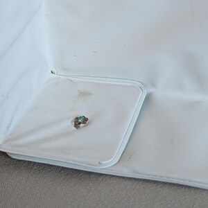 May include: Close-up of a light blue fabric with a square pocket. The pocket has a light blue border and a small, tarnished metal detail with a turquoise center. The fabric shows some discoloration.